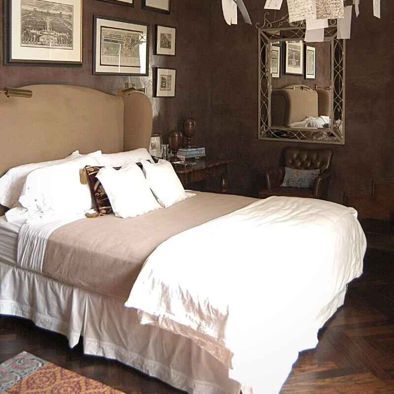 Photograph of a guest room with suede brown walls, with vintage drawings hanging. The bed has an apolstered headboard and is made with a tan duvet and pillows. There is a leather chair under a mirror in the corner. A room idea for when the kids move out.