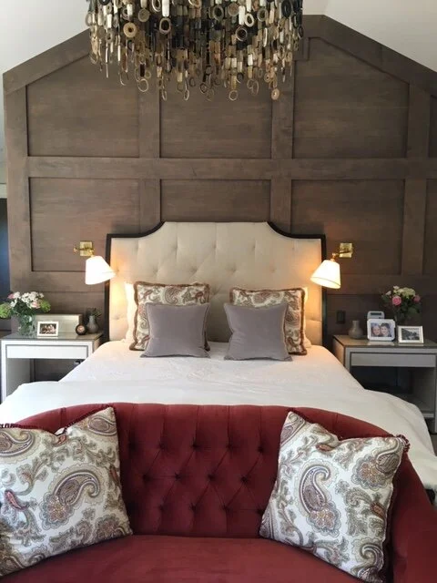Bedroom with an upholstered headboard.  The bed is flanked by reading lights and the wall behind the bed is geometric dark wood.  In front of the bed is a red couch with throw cushions.  A modern metal chandelier hangs above.  This complements an article about choosing mattresses.