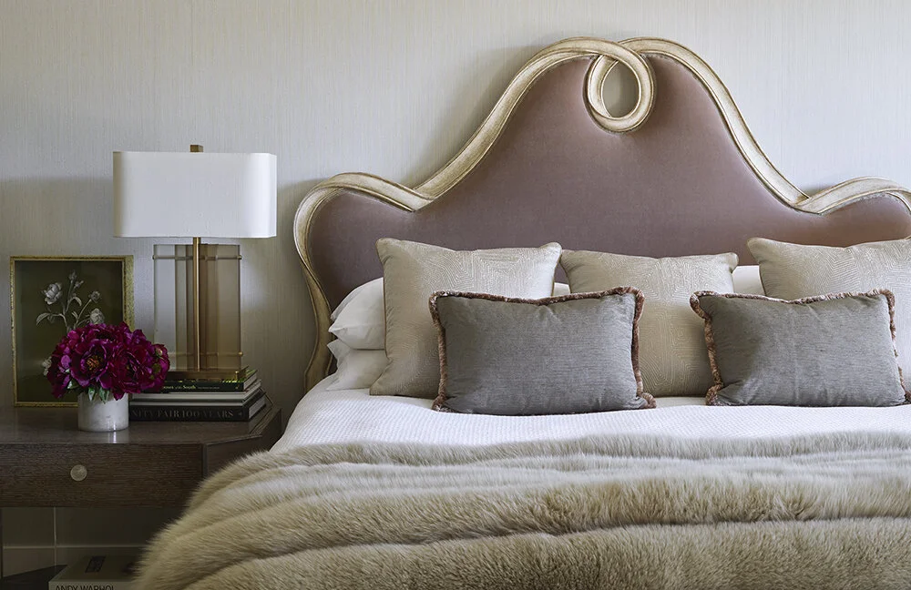 A close up of a bed and nightstand.  The bed is appointed with a fuzzy blanket and throw pillows against an upholstered headboard.  The side table has a modern rectangular lamp and a short vase of burgundy flowers.