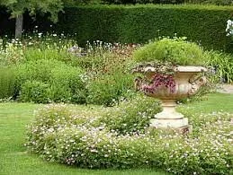 A vintage urn contains plants draping over it in the middle of a garden space.