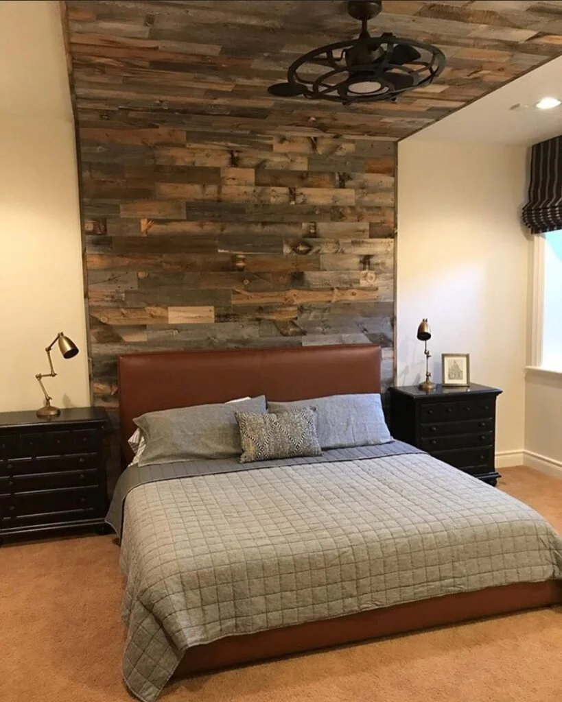 This bedroom is created a round a Feature Wall made of wood planks.  The bed is covered with a grey duvet and grey pillows, accented by  a deep brown headboard and black side tables.