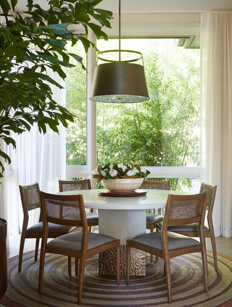 A small dining room with a white circular table topped with a flower arrangement.  A plant is in the foreground.