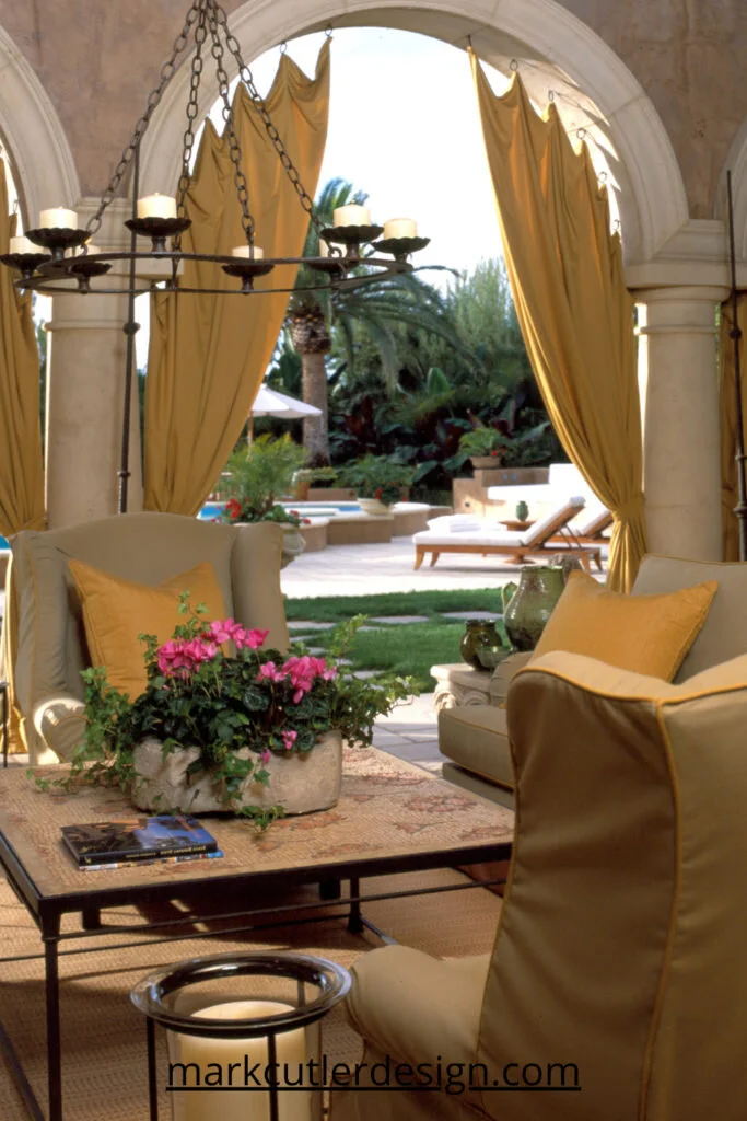 This Loggia in an Italian style home in Beverly Hills is accented with drapes that help to create a sense of enclosure and the custom outdoor furniture modelled on indoor antique pieces make this traditional style room feel sumptuous. An antique tile panel was repurposed as a coffee table and the candle lit chandelier makes for a romantic feeling.