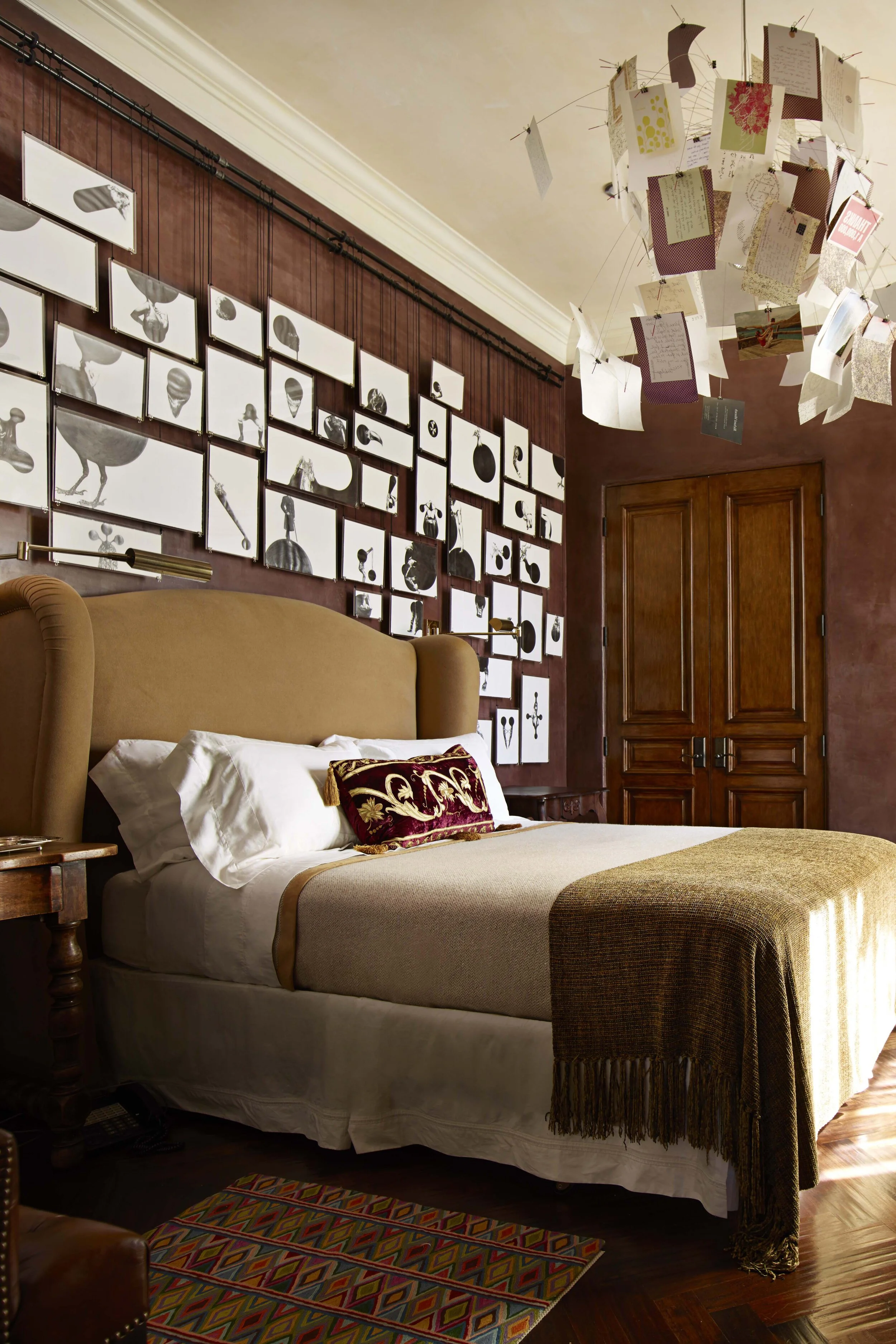 A lush bedroom with dark brown patina walls.  Art is hung gallery style behind the bed.  The chandelier is made of paper ephemera.  Rehanging art is a great way to do a holiday refresh.