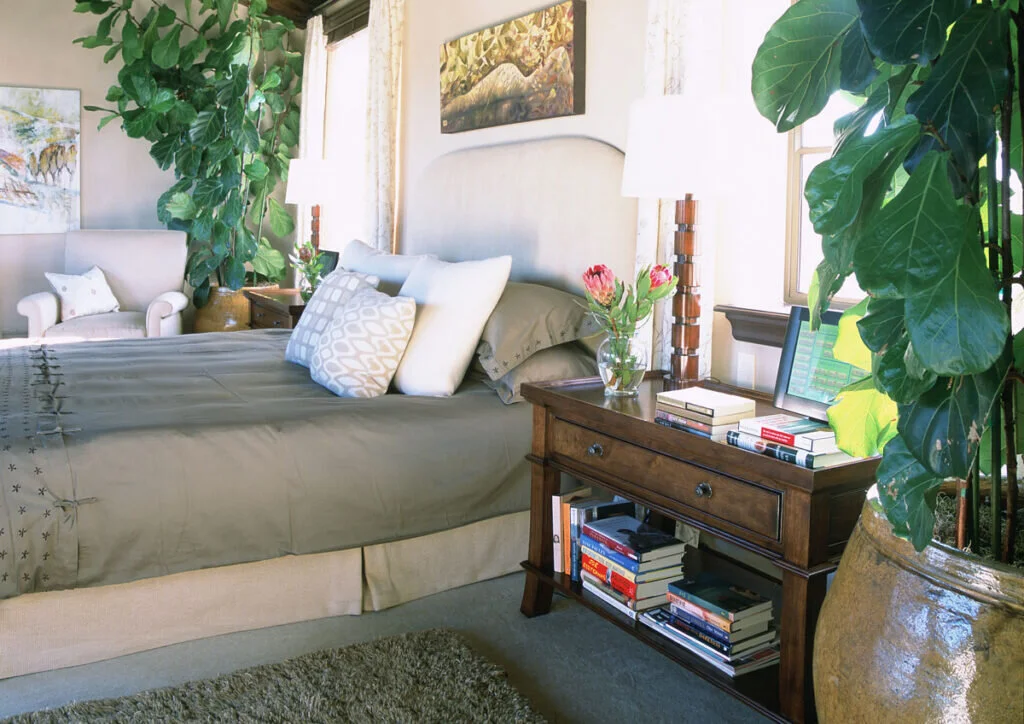 Natural light and lamps provide a lovely, cozy atmosphere for this bedroom. The bed is flanked by a side table with books and two large green plants.
