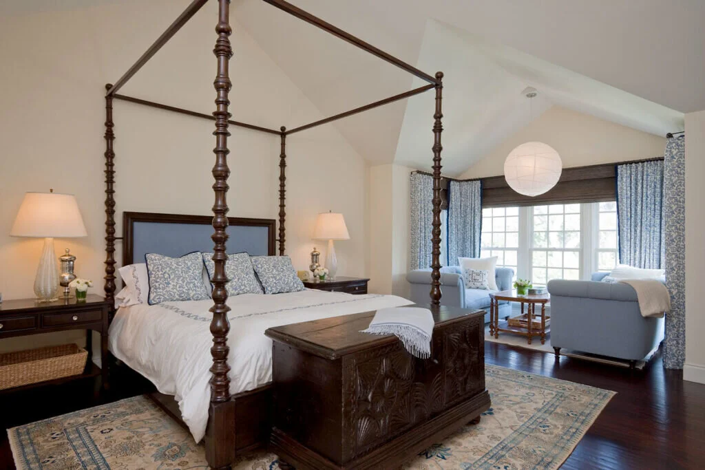A cozy bedroom with a large four poster canopy style bed accented with blue. A seating area contains two comfortable armchairs in a window nook.